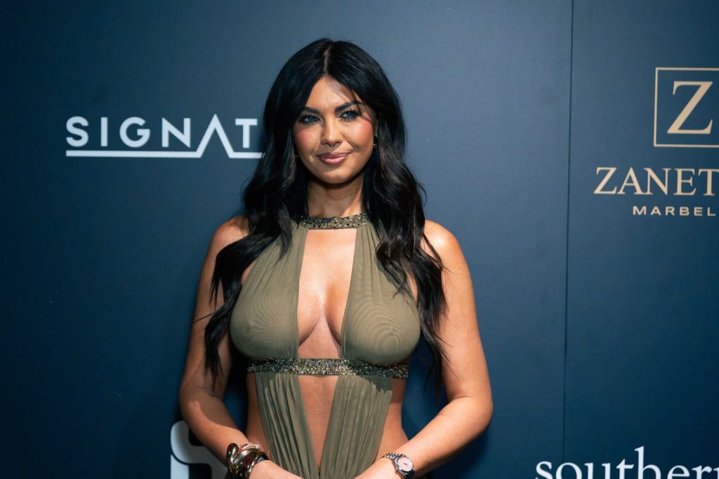 Woman with long dark hair wearing a sleeveless, olive-green, cut-out dress stands in front of a dark blue backdrop with white and gold logos. She is smiling slightly and has her hands in front of her.