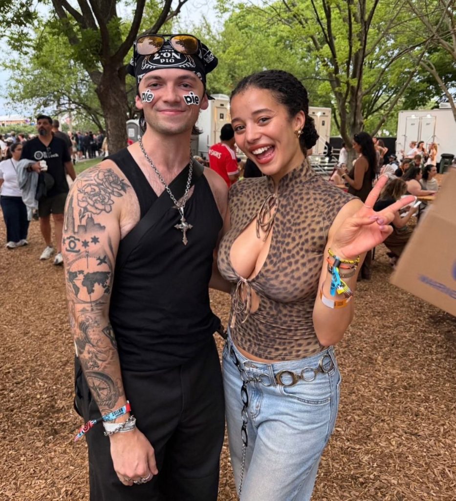 Two people pose outdoors at a festival. The man wears a black bandana, sunglasses, and a black top with tattoos visible. The woman wears a leopard print halter top and jeans, smiling and making a peace sign. Trees and people are in the background.