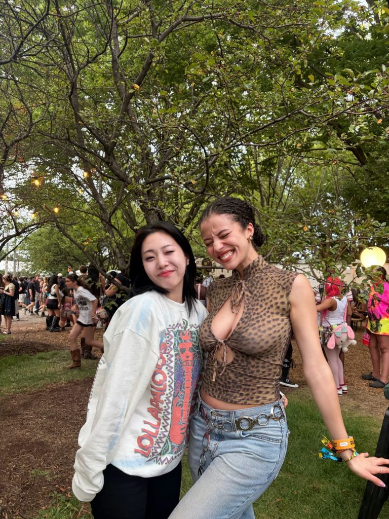 Two women smile while standing outdoors at a festival. One wears a graphic sweatshirt, the other a leopard print sleeveless top and jeans. People and trees are in the background.