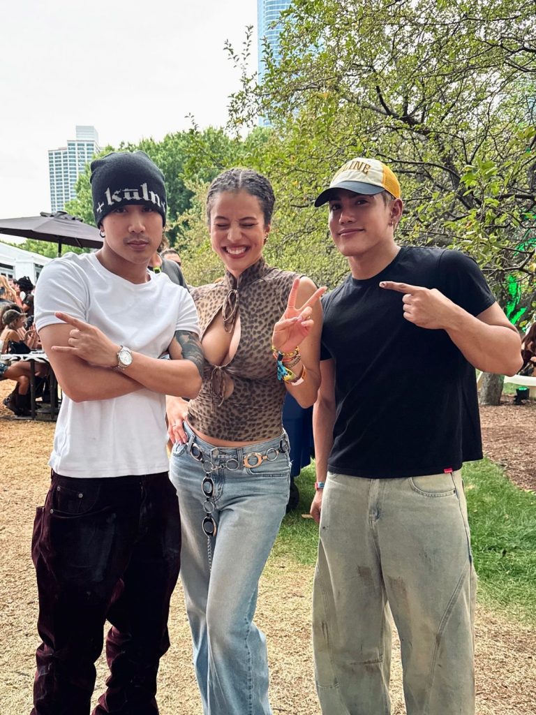 Three people pose outdoors at a festival; the woman in the center smiles and flashes a peace sign, flanked by two men, one crossing his arms and the other pointing at her. Trees and people can be seen in the background.