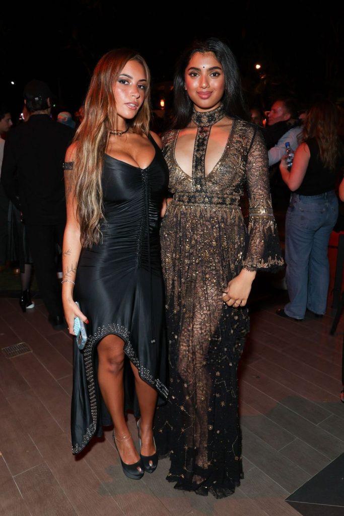 Two women posing together at a nighttime event; one wears a black fitted dress with off-the-shoulder straps, the other wears a long, sheer, sparkly dress with long sleeves and a keyhole neckline. People and lights are in the background.