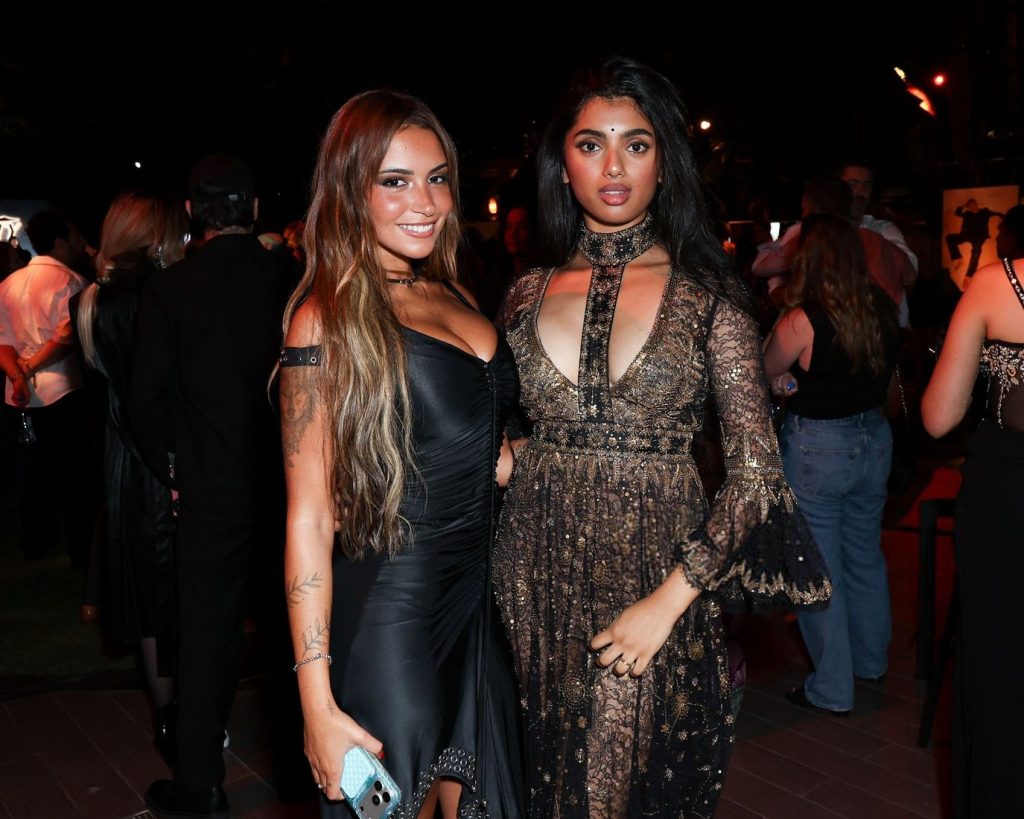 Two women pose together at a nighttime event. One wears a black dress and holds a phone, while the other wears a detailed, long-sleeved lace gown. A crowd of people mingles in the background under dim lighting.