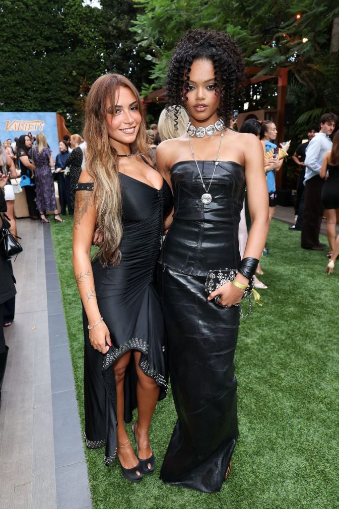 Two women in black dresses pose together at an outdoor event on grass, surrounded by other guests. One has long light brown hair, the other has short curly hair and wears a choker necklace.