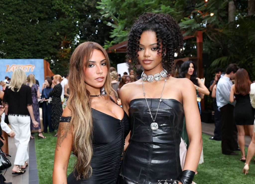 Two women in black dresses pose outdoors at an event. One has long brown hair and tattoos, the other has curly hair and wears a silver choker. People are mingling in the background.