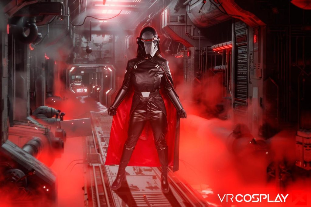 Woman in full black helmet and black leather bodysuit with red cape standing in a red-lit sci-fi corridor filled with smoke.