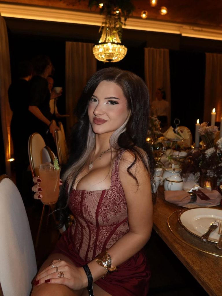 A woman in a maroon lace dress sits at a decorated dinner table, holding a drink and smiling. The setting is elegant with candles, floral arrangements, and a chandelier above.
