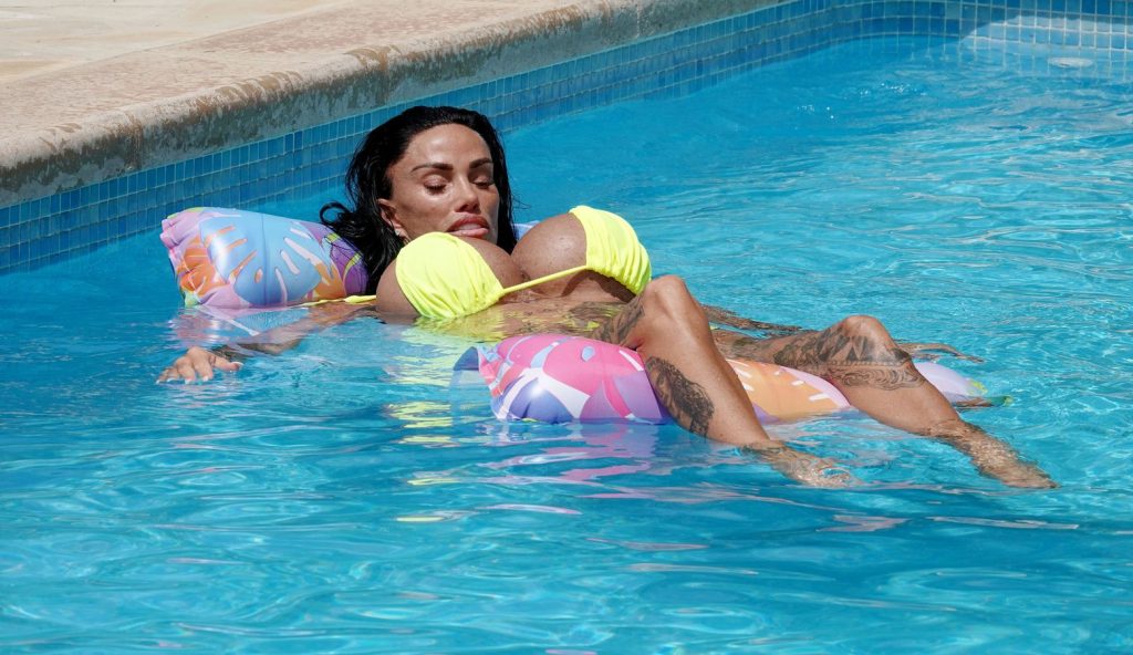 A woman with long dark hair in a yellow bikini relaxes on a colorful pool float in a swimming pool, eyes closed and legs stretched out.