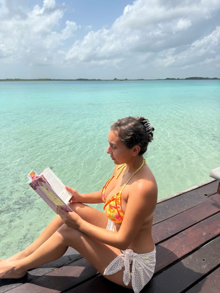 A woman in a bikini and white cover-up sits on a wooden dock by clear turquoise water, reading a book under a partly cloudy sky.