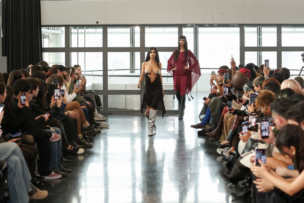 Two models walk down a runway during a fashion show, surrounded by an audience taking photos with their phones in a bright, modern room with large windows.