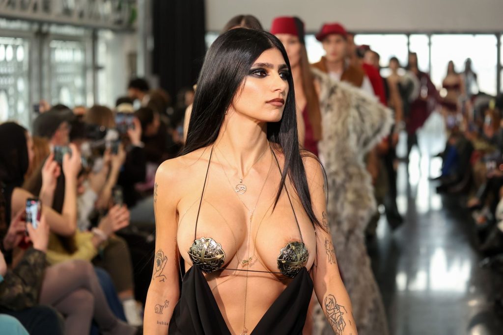 A model with long black hair walks a runway wearing a revealing black dress with metallic leaf-shaped pasties, as an audience takes photos and other models follow behind.