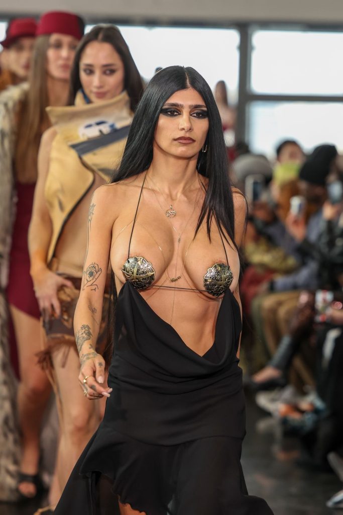 A model with long black hair walks a fashion runway wearing a revealing black dress with metallic leaf nipple covers. Other models and an audience are seen in the background.