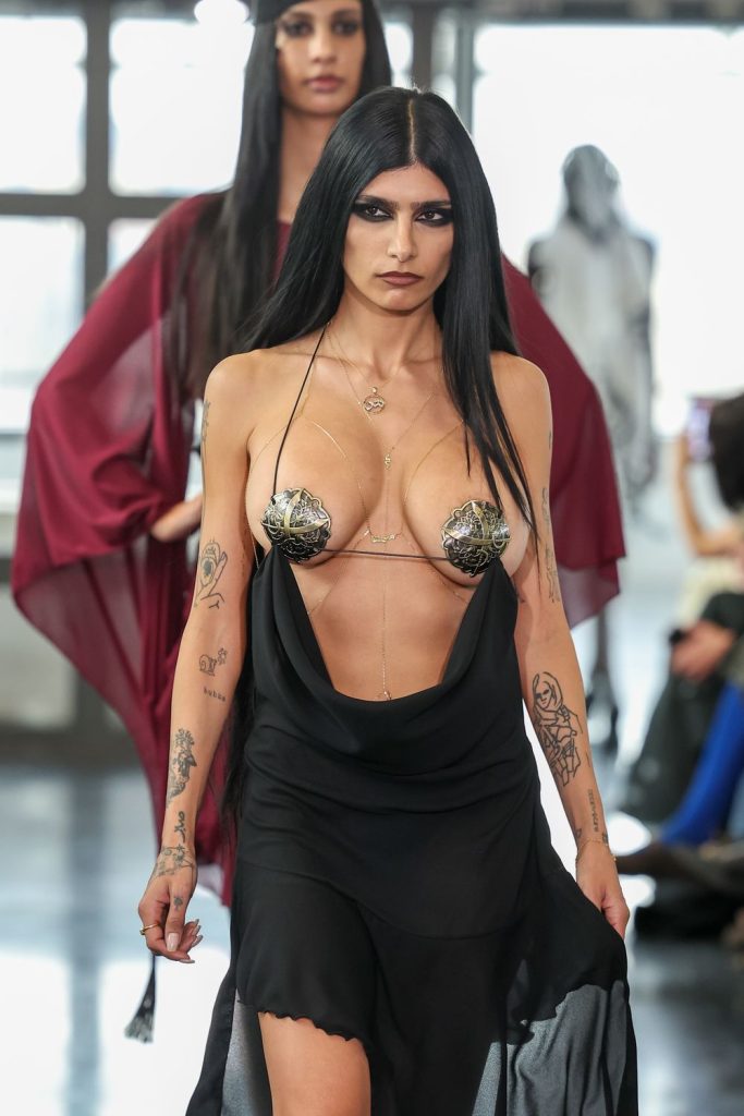 A model with long black hair walks the runway in a revealing black dress with metallic embellishments, displaying tattoos on her arms. Another model in a burgundy outfit follows behind.