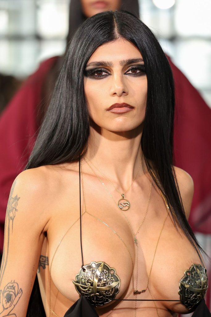 A woman with long black hair and dramatic eye makeup wears a revealing black and gold top, gold necklaces, and has visible tattoos on her arms. The background is blurred, suggesting a fashion event or runway.