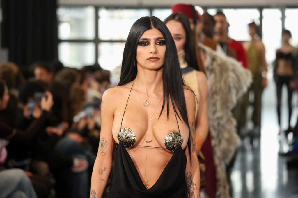 A model with long black hair, bold eye makeup, and tattoos walks a runway wearing a black dress with ornate gold nipple covers. An audience watches in the background.