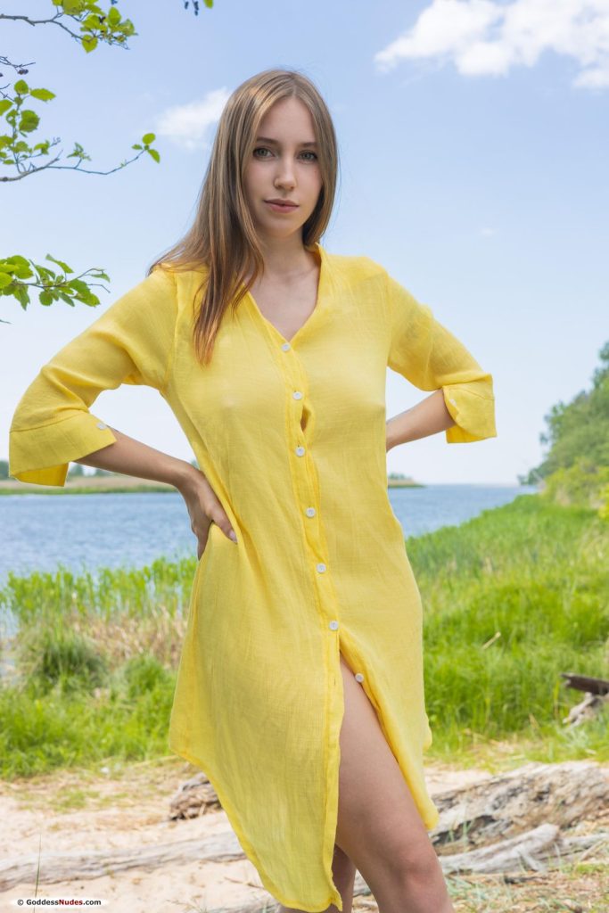 Woman with long light brown hair in a yellow button-up shirt dress standing outdoors by a lake with hands on her hips.