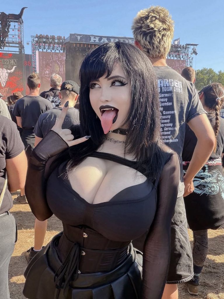 A person in a black gothic outfit and mask poses at an outdoor concert, making a rock hand gesture and sticking out their tongue. People in dark clothing stand around, and a stage is visible in the background.