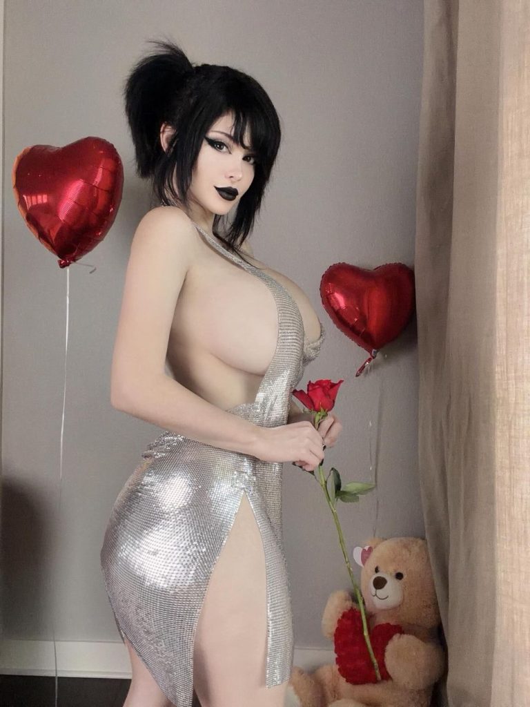 A woman with dark hair in a silver dress holds a red rose, standing next to a teddy bear and heart-shaped balloons in a room.