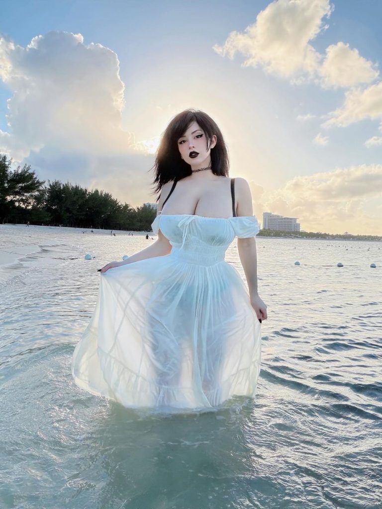 Woman in a sheer white dress stands in shallow ocean water at sunset, with sunlight creating a glowing effect behind her. Trees and a tall building are visible in the background.