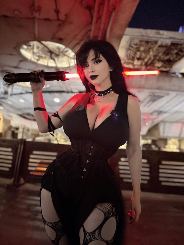 A woman in a black gothic outfit with cut-out leggings holds a red lightsaber over her shoulder, posing in a sci-fi setting with industrial details and dramatic lighting.