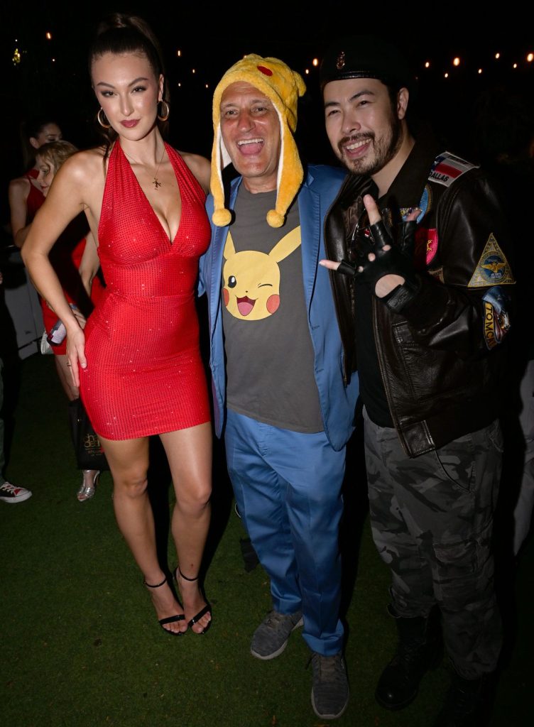 Three people pose together at a nighttime outdoor event. One wears a red dress, another wears a Pikachu hat and blue suit, and the third wears a black jacket, beret, and camouflage pants. String lights hang in the background.