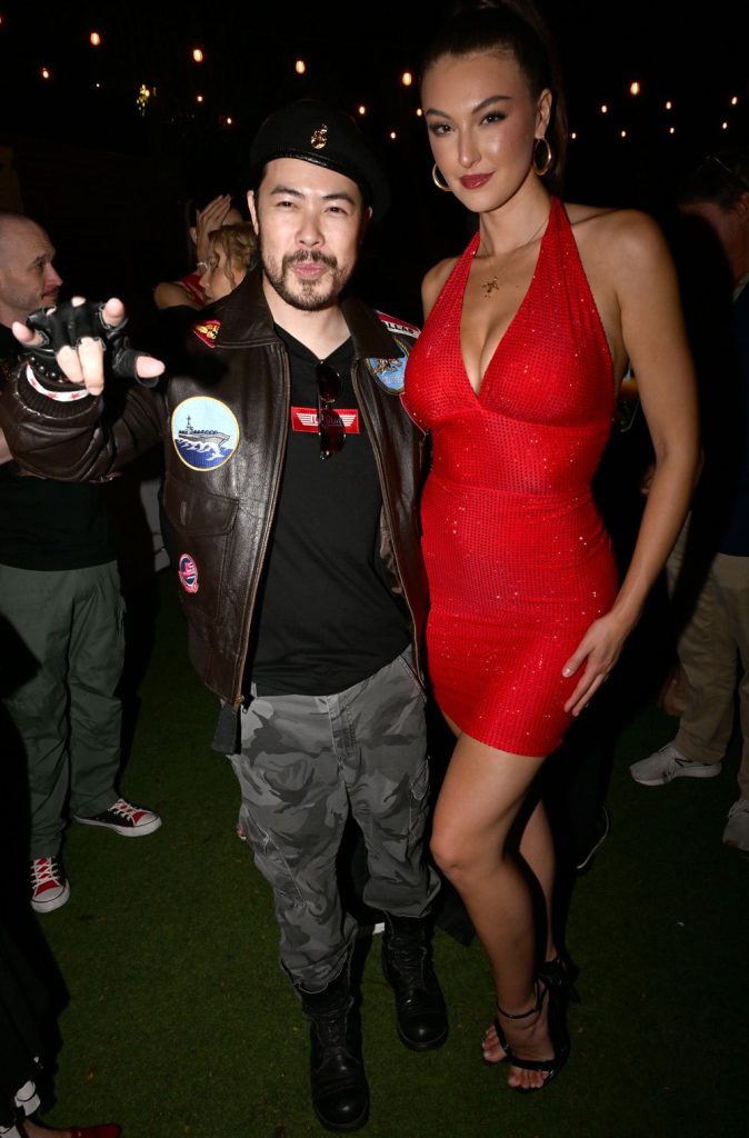 A person in a brown leather jacket and beret poses next to a person in a red sleeveless dress, both smiling at a nighttime outdoor event with string lights and people in the background.