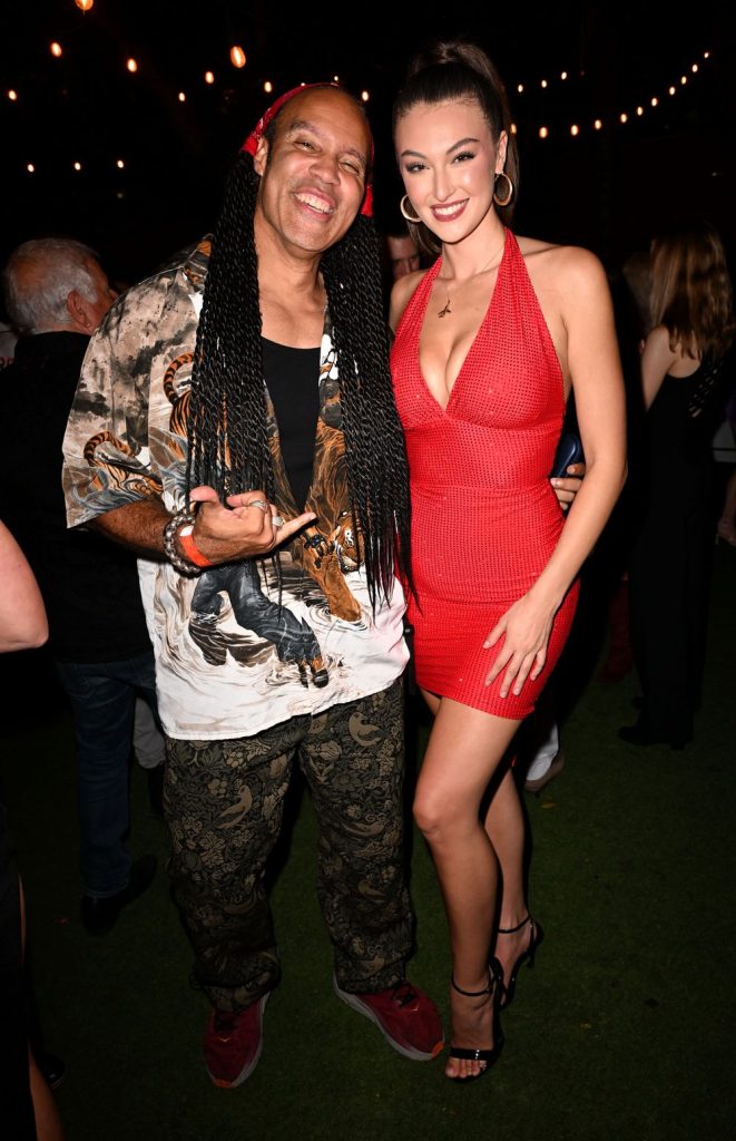 A man with long braids and a woman in a red dress smile and pose together at an outdoor event at night, surrounded by other people.