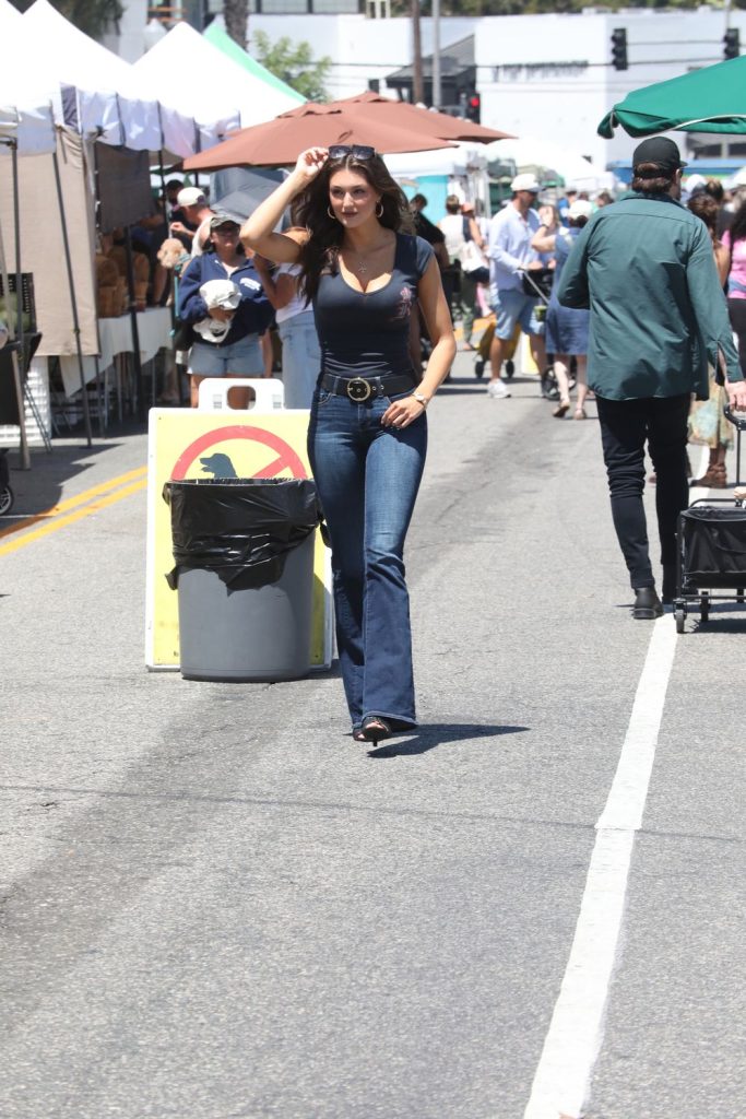 A woman in a blue outfit and sunglasses walks down a market street lined with tents and people, passing a trash can with a no smoking sign.