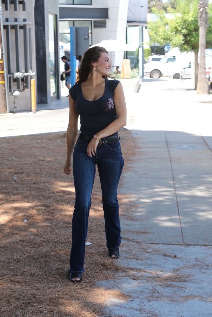 A woman with long brown hair, wearing a navy blue top and dark jeans, walks on a sidewalk outdoors, smiling and looking to her right. Trees and buildings are visible in the background.