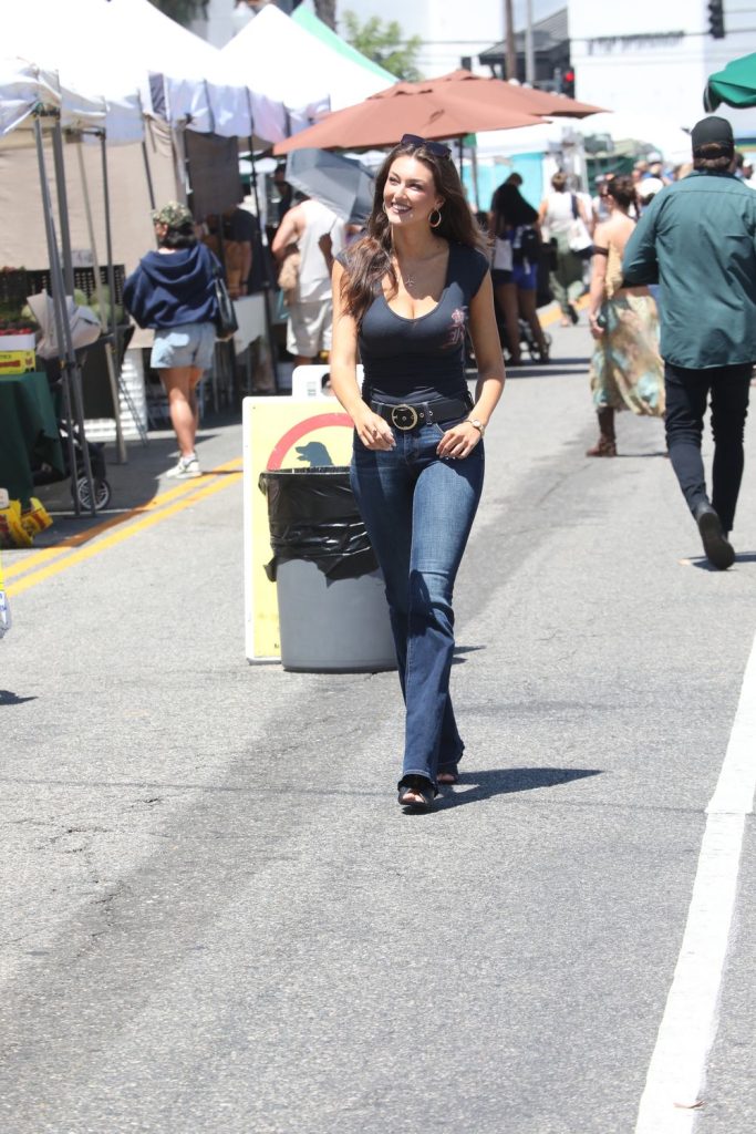 A woman in a black t-shirt and jeans walks and smiles at an outdoor market, with tents, people, and booths visible in the background on a sunny day.