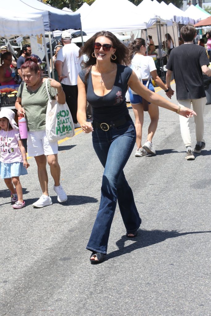 A woman in dark sunglasses, a black top, and jeans smiles while walking at an outdoor market. Other people, tents, and stalls are visible in the background on a sunny day.