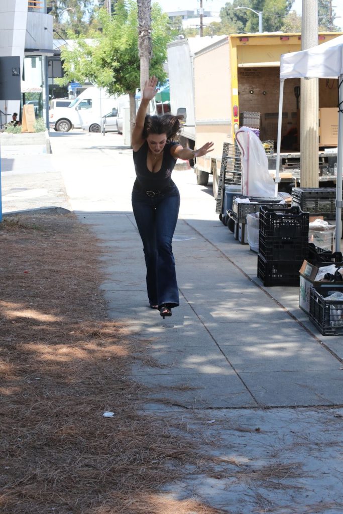 A woman in a black top and jeans balances on one foot with arms raised, walking along a sidewalk next to market stalls with crates and boxes in daylight.