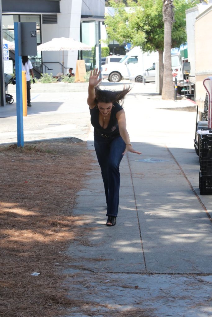 A woman in dark clothes, with her arms extended forward and one leg up, appears to be balancing or playfully posing on a sidewalk outdoors, with trees and vehicles in the background.