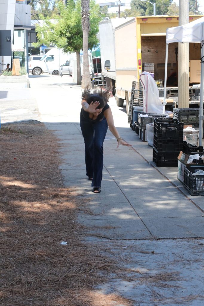 A person with long dark hair, wearing a black top and jeans, leans forward on a sidewalk with one arm extended and the other hand covering their face, near plastic crates and trucks.