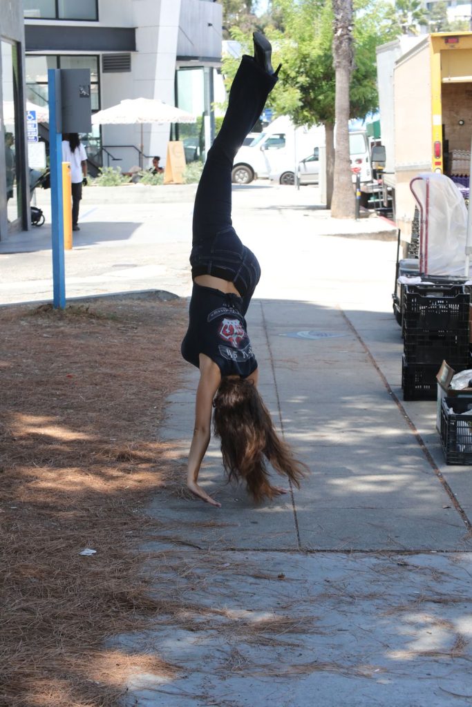 A person with long hair does a cartwheel on a sidewalk near trees, crates, and buildings on a sunny day.