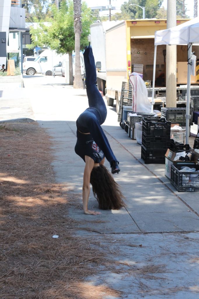 A woman with long hair performs a handstand on a sidewalk near market stalls and plastic crates, with trees and vehicles in the background.