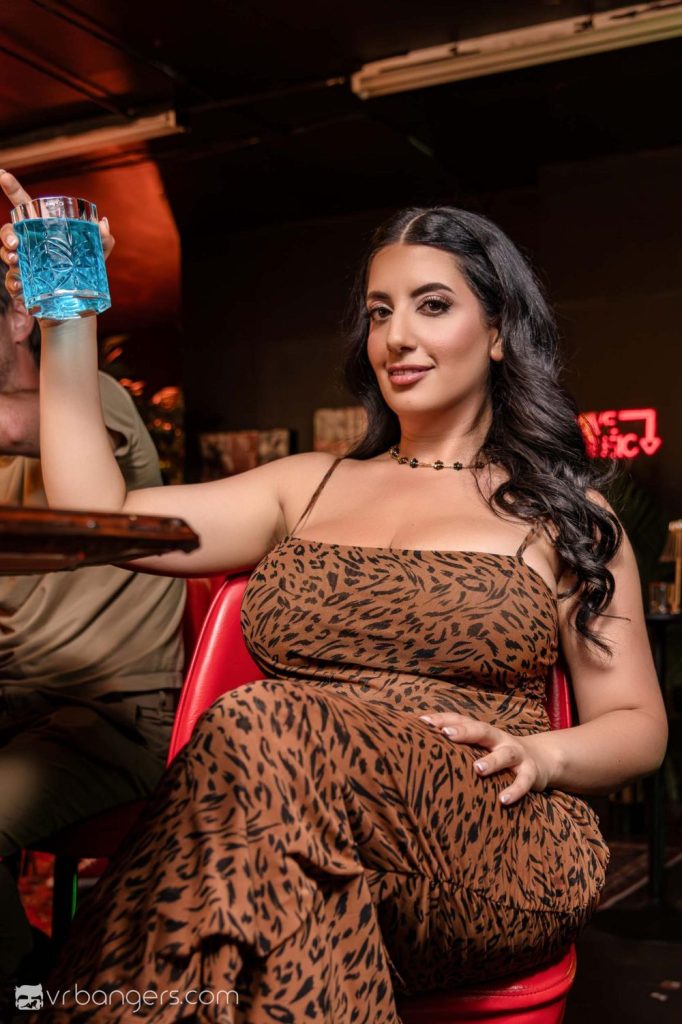 A woman in a brown animal print dress sits on a red chair, holding up a blue drink and smiling at the camera in a dimly lit bar or lounge.