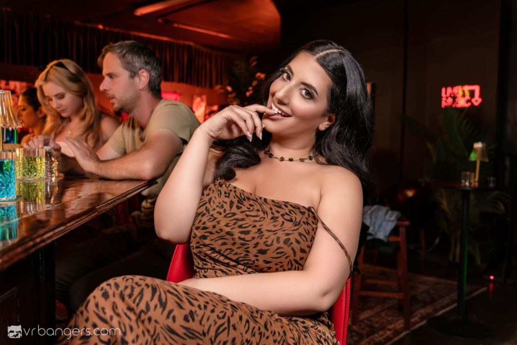 A woman in a leopard-print dress sits at a bar, smiling and touching her lip. Two people talk in the background. The setting is dimly lit with colorful drinks on the bar and a neon sign on the wall.