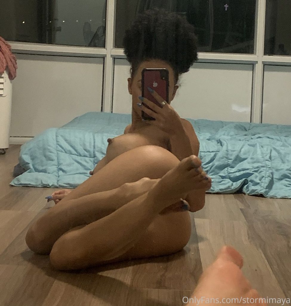 Nude black girl with afro hair sit on her side with feets raised on the floor while taking a selfie.