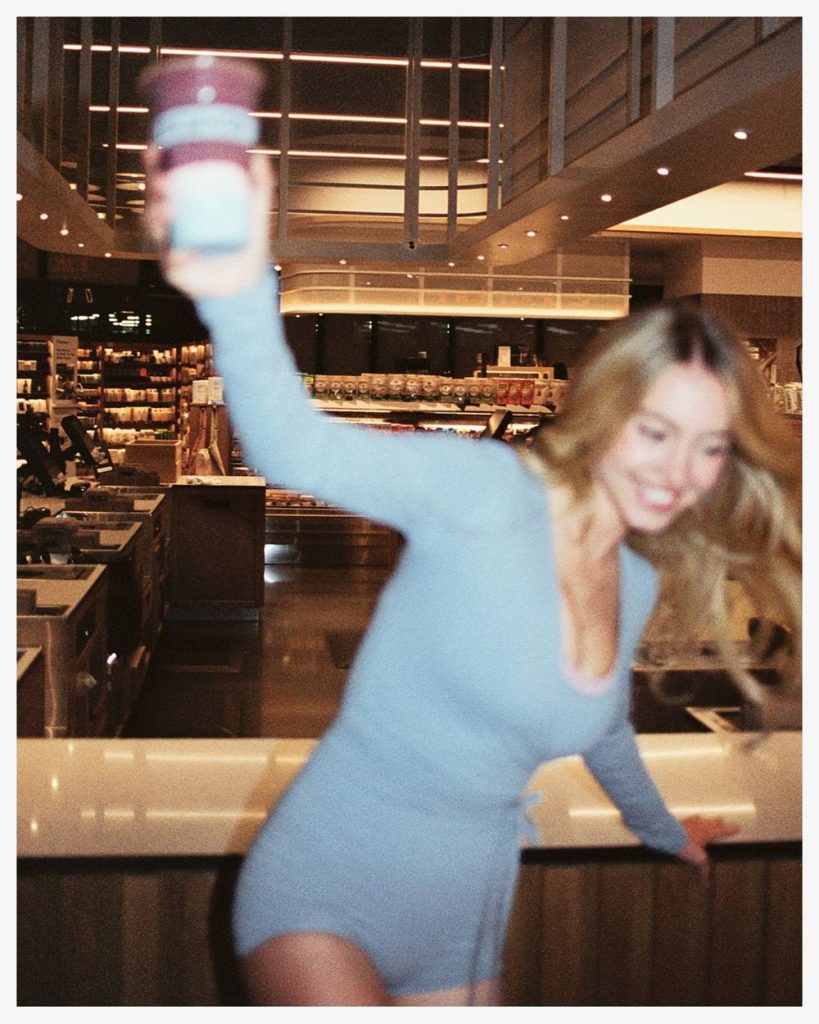 A woman in a light blue outfit smiles and holds up a container in a brightly lit store, with shelves and counters visible in the background. The image is slightly blurred, suggesting motion.