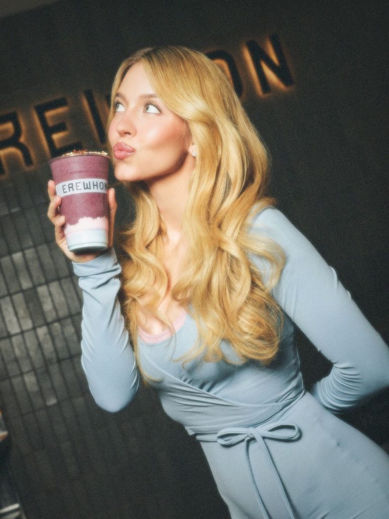 A woman with long blonde hair in a light blue dress holds a purple smoothie and poses with puckered lips. The word EREWHON is visible in the background.