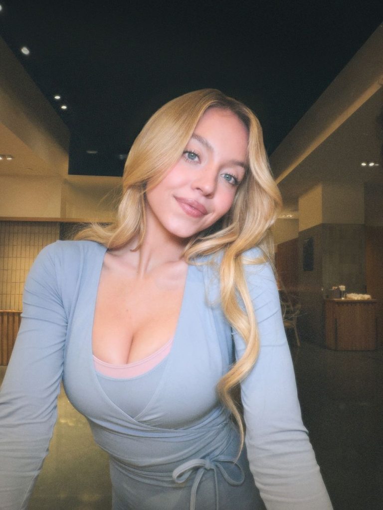 A woman with long blonde hair, wearing a light blue wrap dress, poses smiling indoors in a softly lit, modern room with a dark ceiling and wooden accents.