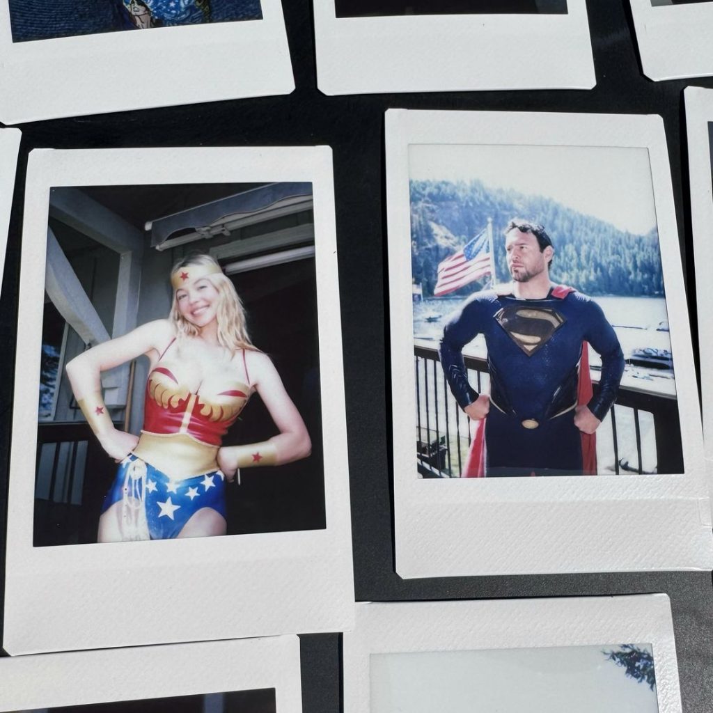 Two Polaroid photos: one shows a woman dressed as Wonder Woman posing with hands on hips; the other shows a man dressed as Superman posing with hands on hips, an American flag and lake in the background.