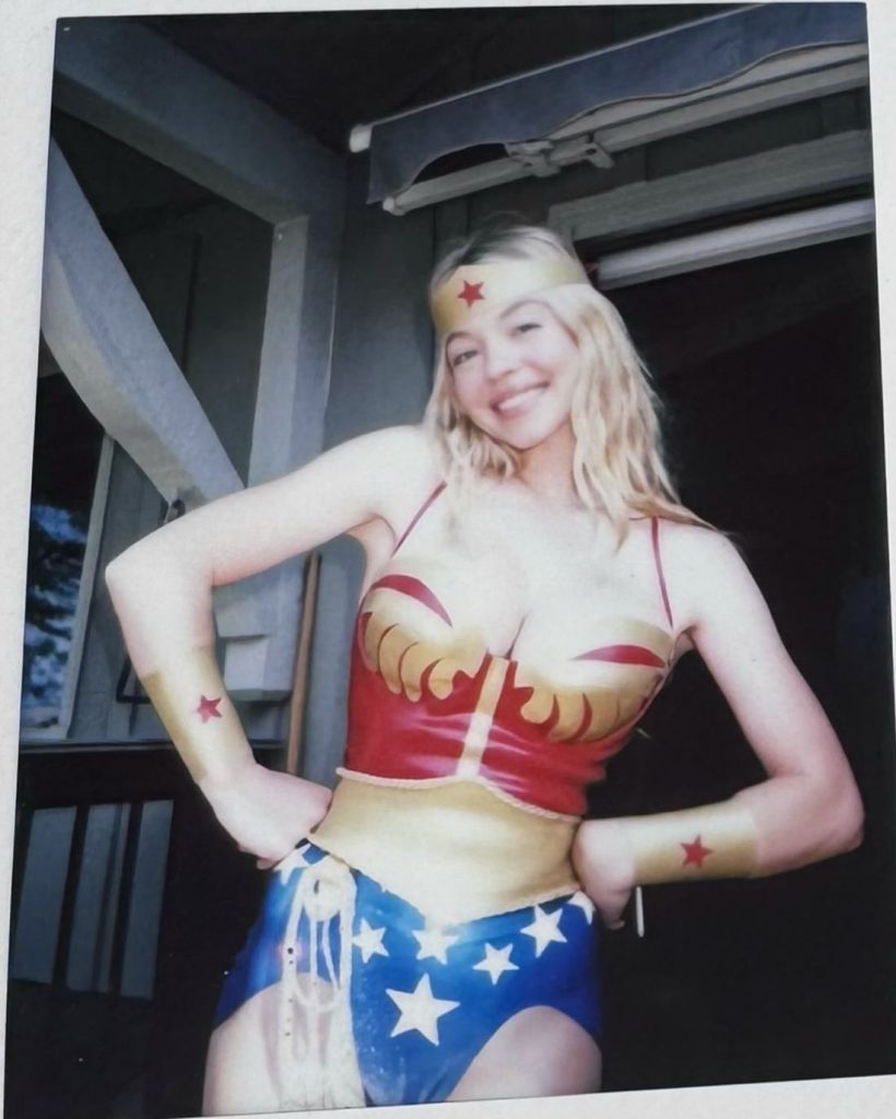 A smiling woman poses in a Wonder Woman costume, featuring a red and gold top, blue shorts with white stars, gold belt, headband, and wrist cuffs, standing in front of a building.