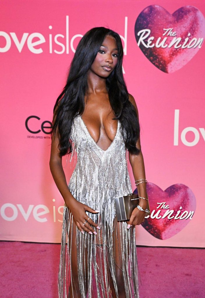 A woman in a silver fringed dress poses on a pink carpet at a Love Island: The Reunion event, standing in front of a pink backdrop with show logos.