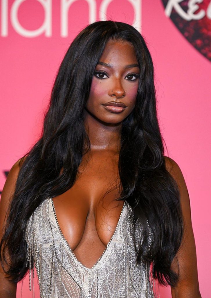 A woman with long black hair and a silver, low-cut dress poses in front of a pink background at an event.