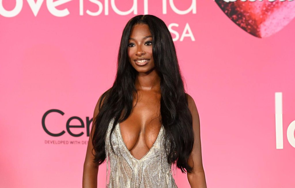 A woman with long black hair in a low-cut silver dress poses on a pink carpet in front of a Love Island USA backdrop.