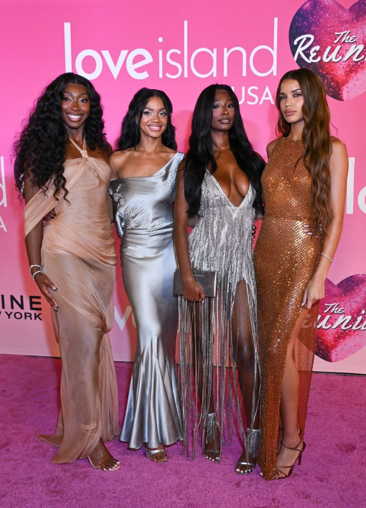 Four women dressed in glamorous evening gowns stand together, smiling, on a pink carpet at a Love Island USA: The Reunion event, with a pink backdrop featuring show logos.