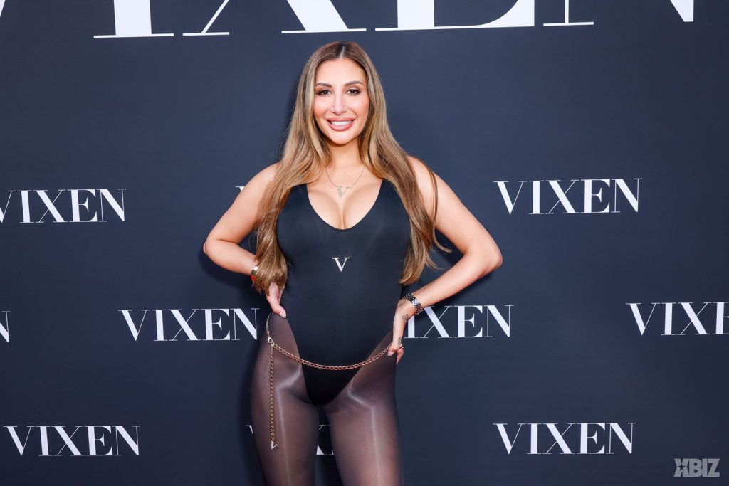 A woman with long light brown hair, wearing a black bodysuit and sheer brown leggings, stands smiling in front of a VIXEN-branded backdrop at an event.
