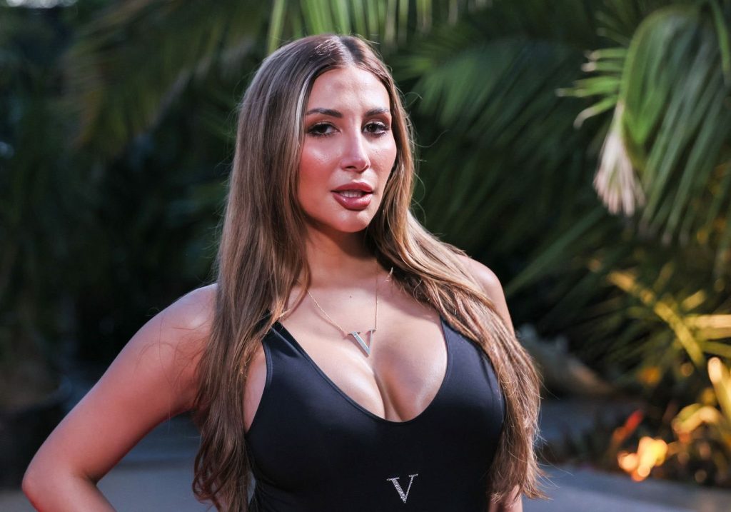 A woman with long brown hair wearing a black sleeveless top and a necklace with a V pendant, stands outdoors in front of green tropical plants.