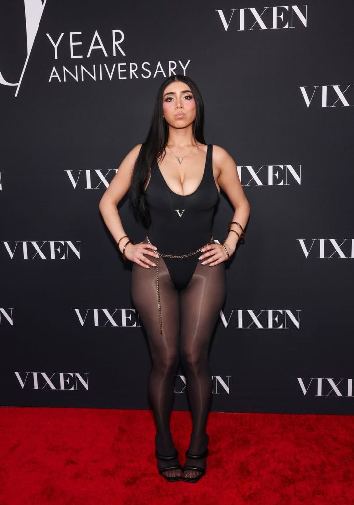 A woman with long black hair in a black bodysuit, sheer tights, heels, and gold accessories stands on a red carpet in front of a backdrop reading VIXEN and YEAR ANNIVERSARY..